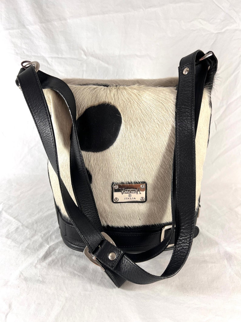 Puede incluir: Un bolso de cubo de piel de vaca blanco y negro con una correa de cuero negro y una hebilla. El bolso tiene una placa de metal plateado con el texto "VALENTINA ITALIA" en ella.