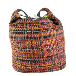 May include: A bucket bag with a woven pattern in pink, orange, green, and yellow. It has black handles and gold-coloured details. The bag is made of a woven material, and is a stylish accessory.