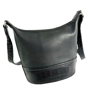 May include: Black leather shoulder bag with a braided strap. The bag has a cylindrical shape with a textured bottom section. The strap is attached with silver-colored hardware.
