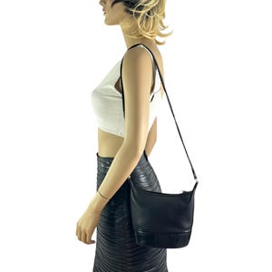 May include: Black leather shoulder bag with a rectangular shape and a long, thin strap. The bag has a textured bottom panel and a zipper closure. The bag is worn over the shoulder.