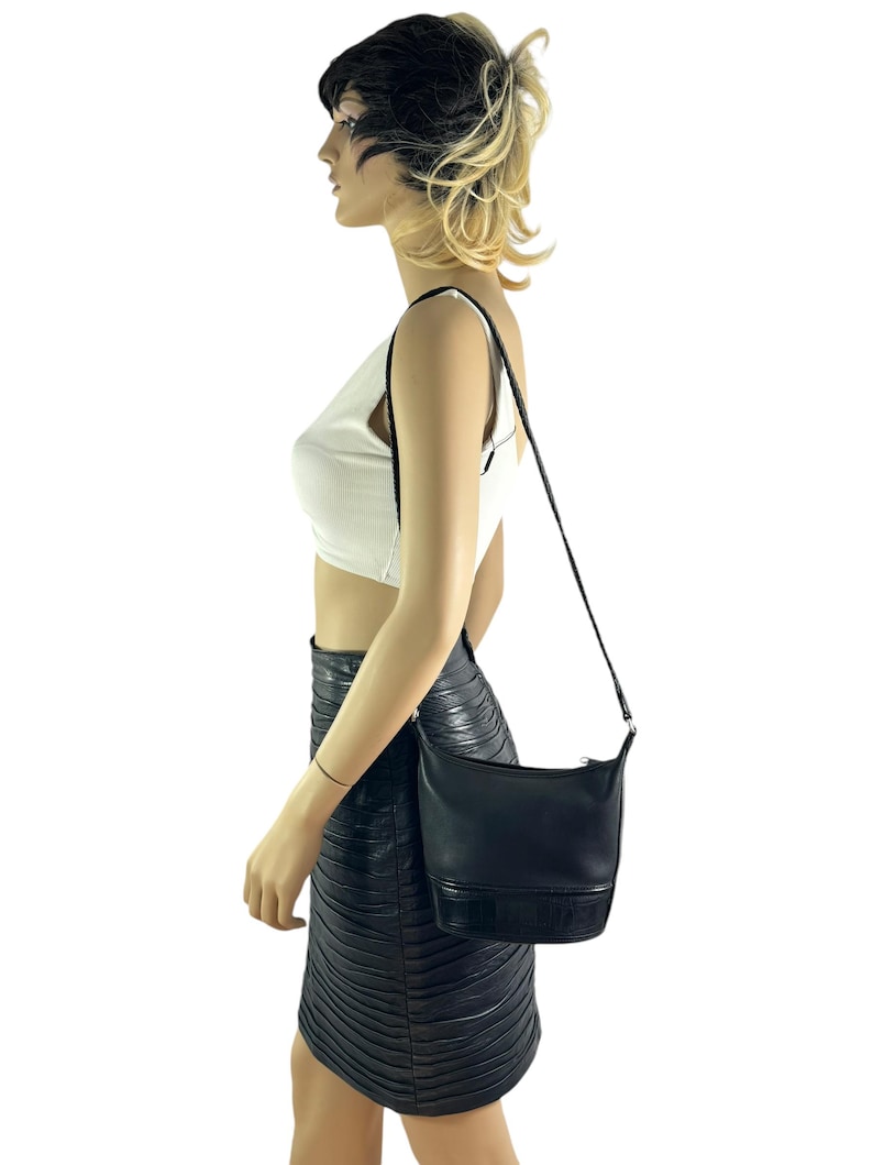 May include: Black leather shoulder bag with a rectangular shape and a long, thin strap. The bag has a textured bottom panel and a zipper closure. The bag is worn over the shoulder.