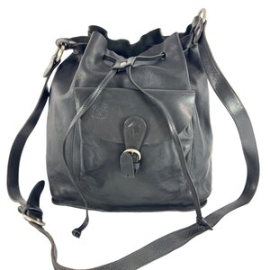 May include: A black leather bucket bag with a drawstring closure and a long adjustable strap. The bag has a front pocket with a buckle closure.