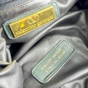 May include: Close-up of the interior of a black leather Valentino Garavani handbag. The bag features a gold-coloured rectangular label with the brand name and logo, and a green leather tag that reads "Made in Italy Genuine Leather."