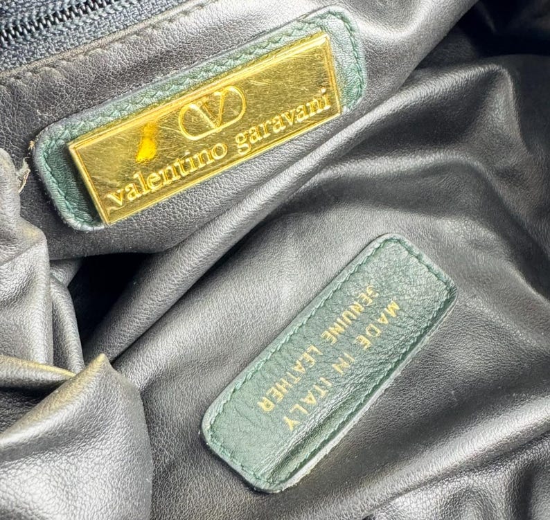 May include: Close-up of the interior of a black leather Valentino Garavani handbag. The bag features a gold-coloured rectangular label with the brand name and logo, and a green leather tag that reads "Made in Italy Genuine Leather."