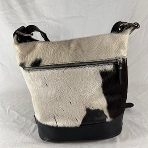 Puede incluir: Un bolso de mano de piel de vaca blanco y negro con una correa y una base de cuero negro. El bolso tiene una cremallera y un peque&ntilde;o bolsillo en la parte delantera.