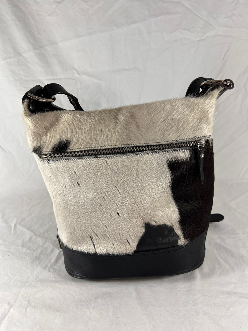 Puede incluir: Un bolso de mano de piel de vaca blanco y negro con una correa y una base de cuero negro. El bolso tiene una cremallera y un peque&ntilde;o bolsillo en la parte delantera.