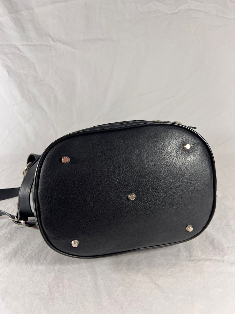 Puede incluir: Bolso de cuero negro de estilo cubo con detalles en metal plateado. El bolso tiene un fondo redondeado y cierre de cremallera.