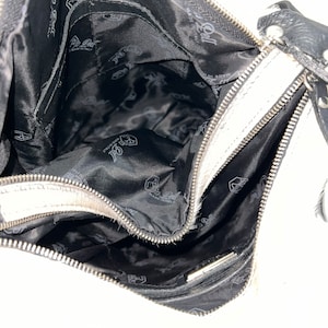 Puede incluir: Bolso de mano de cuero blanco con cremallera negra y forro negro. El forro tiene un logotipo repetido de una mano estilizada sosteniendo un coraz&oacute;n.