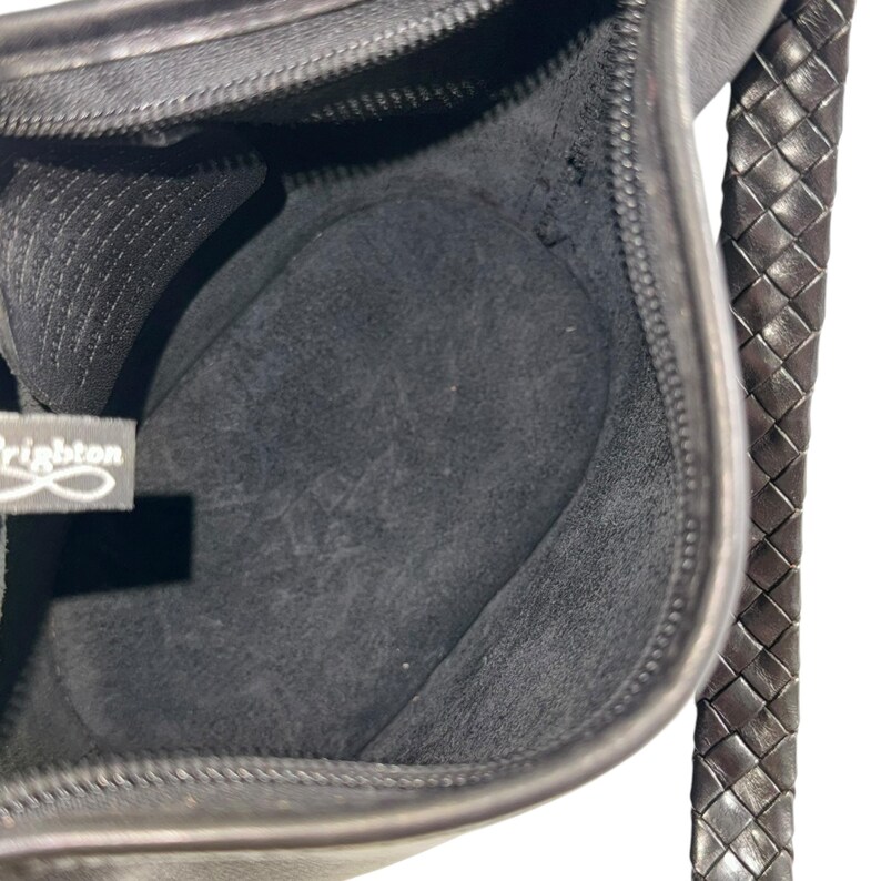 May include: Black leather handbag with a braided strap. The interior of the bag is visible, showing a dark gray lining and a zippered closure. The brand name "Brighton" is visible on a tag inside the bag.