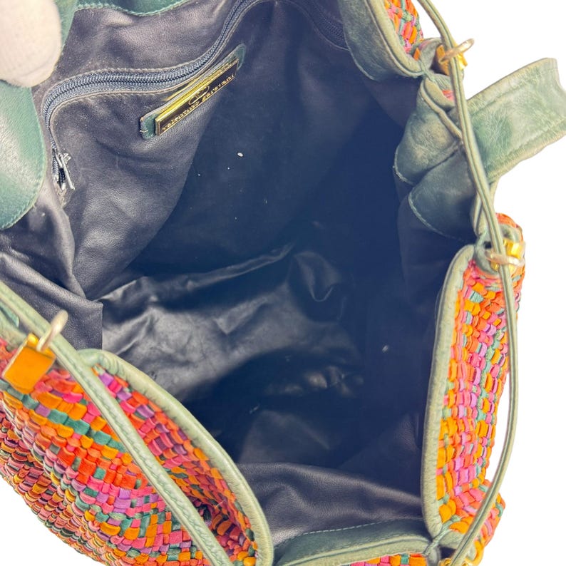 May include: A colourful woven handbag with a drawstring closure. The bag features a vibrant pattern of orange, pink, and purple woven strips. The bag has green leather accents and straps. The interior is black.