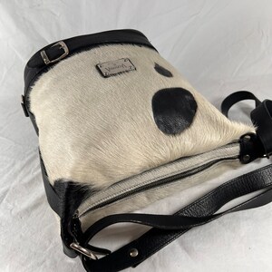 Puede incluir: Un bolso de mano de piel de vaca blanco y negro con una correa de cuero negro y una placa de metal plateado con la palabra "Virginia" en ella.