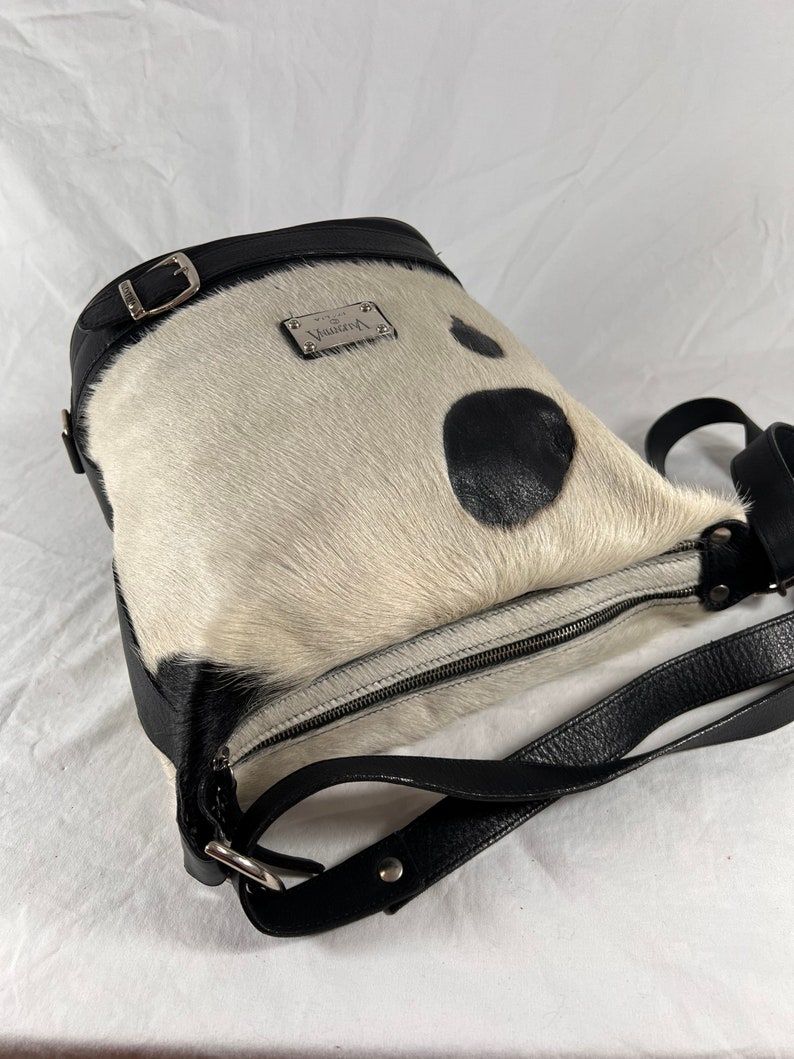 Puede incluir: Un bolso de mano de piel de vaca blanco y negro con una correa de cuero negro y una placa de metal plateado con la palabra "Virginia" en ella.