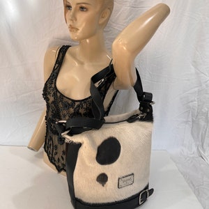 Puede incluir: Un bolso de mano de piel de vaca blanco y negro con una correa de cuero negro y una hebilla. El bolso tiene un parche de cuero negro con el texto "Valentino" en &eacute;l.