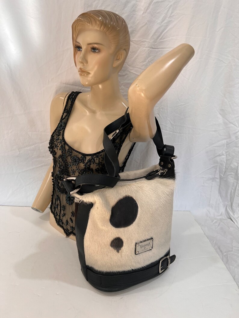 Puede incluir: Un bolso de mano de piel de vaca blanco y negro con una correa de cuero negro y una hebilla. El bolso tiene un parche de cuero negro con el texto "Valentino" en &eacute;l.