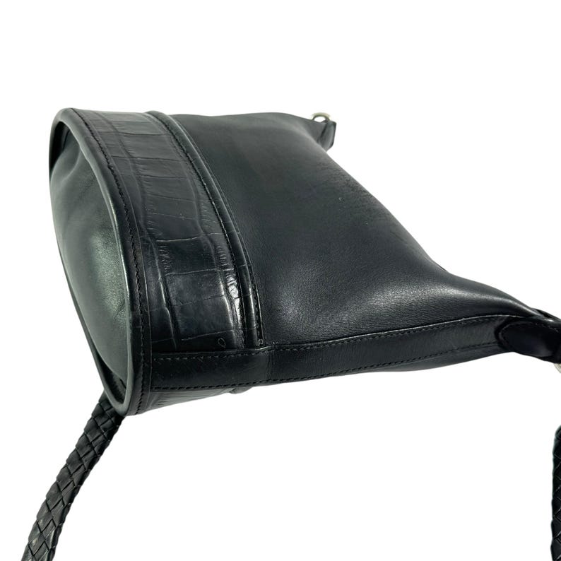 May include: Black leather shoulder bag with a braided strap. The bag features a textured, crocodile-like panel on the front. The bag is angled, showing the top and side.