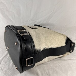 Puede incluir: Un bolso de mano de piel de vaca blanco y negro con una correa de cuero negro y una hebilla plateada. El bolso tiene una placa de metal plateado con la palabra "VALENTINA" en ella.