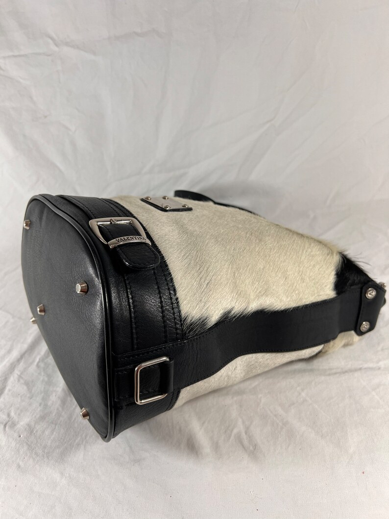 Puede incluir: Un bolso de mano de piel de vaca blanco y negro con una correa de cuero negro y una hebilla plateada. El bolso tiene una placa de metal plateado con la palabra "VALENTINA" en ella.