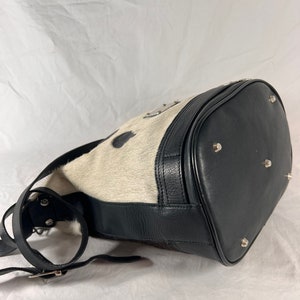 Puede incluir: Un bolso de cubo de piel de vaca blanco y negro con una correa de cuero negro y herrajes plateados. El bolso tiene una forma redondeada y una cremallera.