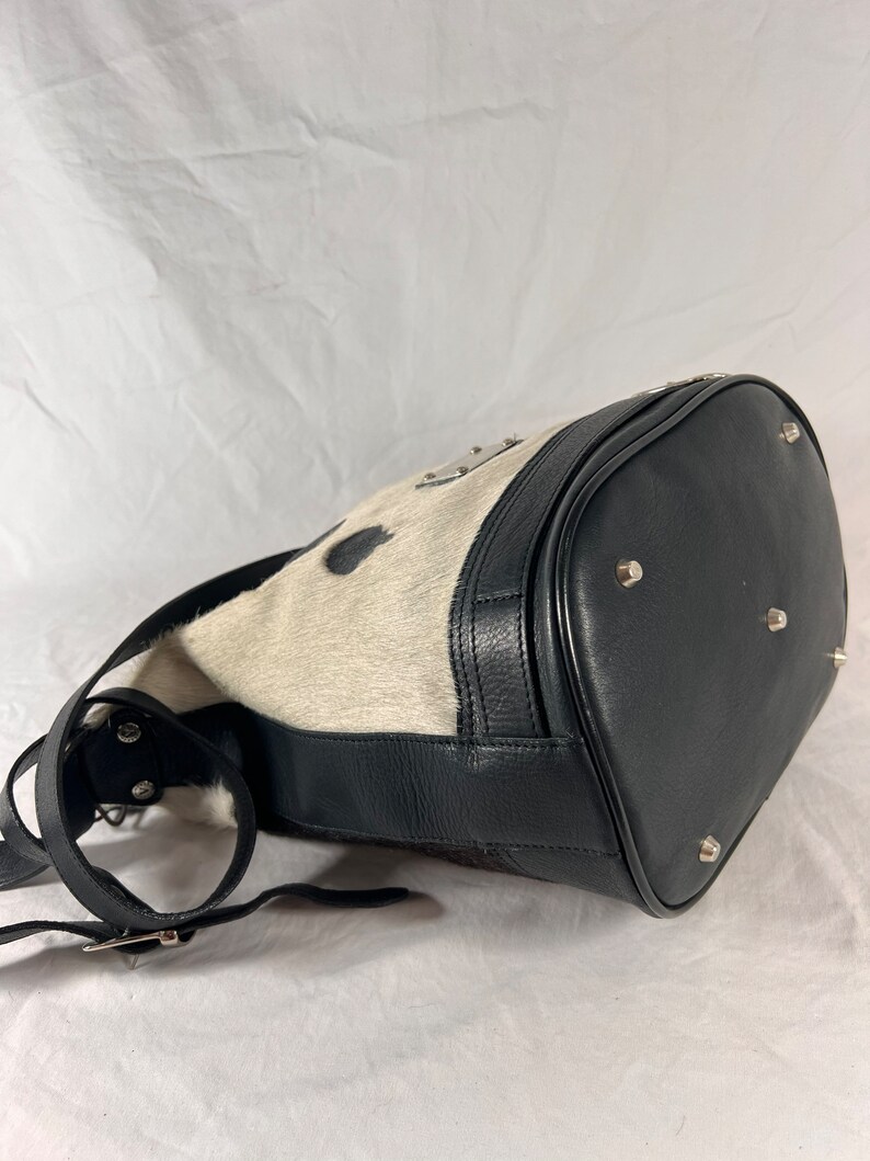 Puede incluir: Un bolso de cubo de piel de vaca blanco y negro con una correa de cuero negro y herrajes plateados. El bolso tiene una forma redondeada y una cremallera.