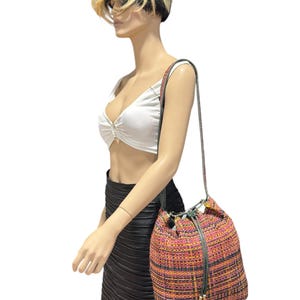 May include: A multi-coloured woven bucket bag with a drawstring closure and a long shoulder strap. The bag features a mix of pink, orange, and yellow threads, creating a textured appearance. The strap is a dark green colour.