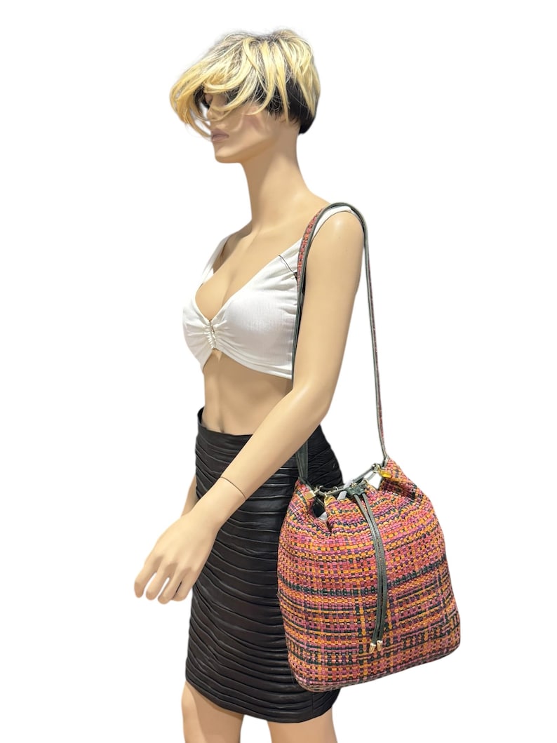 May include: A multi-coloured woven bucket bag with a drawstring closure and a long shoulder strap. The bag features a mix of pink, orange, and yellow threads, creating a textured appearance. The strap is a dark green colour.