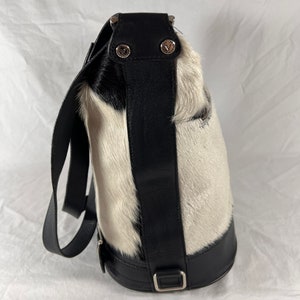 Puede incluir: Un bolso de mano de piel de vaca blanco y negro con una correa y una hebilla de cuero negro. El bolso tiene un asa superior y una correa para el hombro.