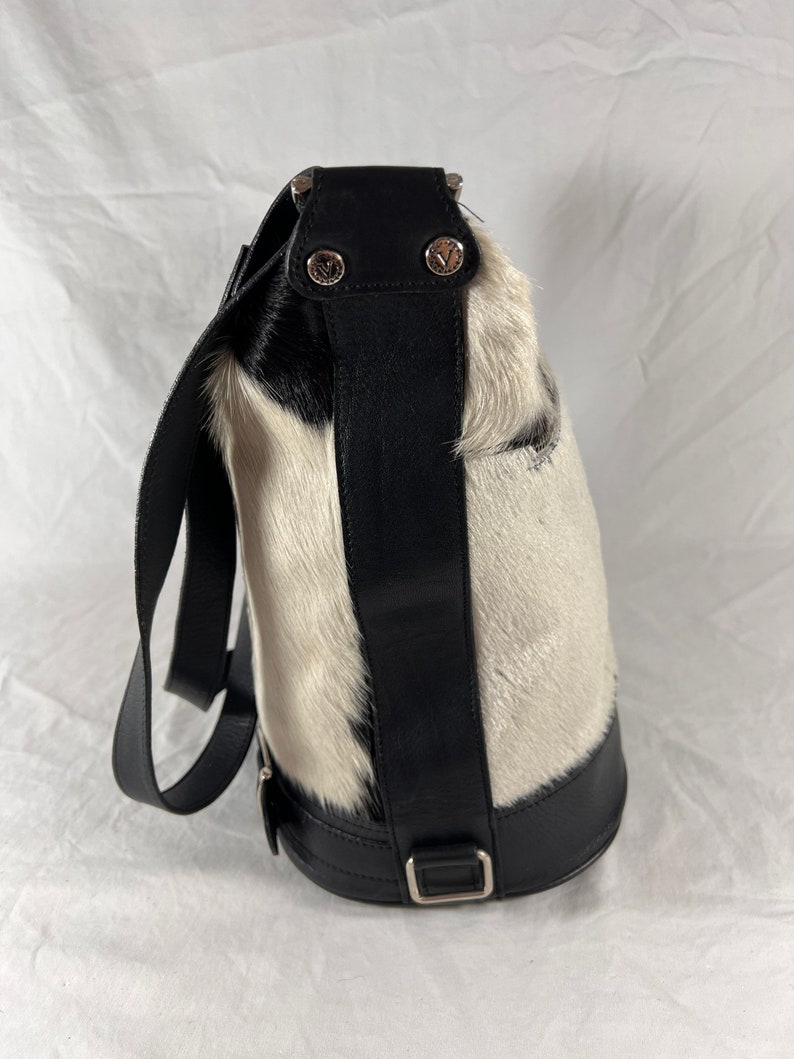 Puede incluir: Un bolso de mano de piel de vaca blanco y negro con una correa y una hebilla de cuero negro. El bolso tiene un asa superior y una correa para el hombro.