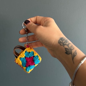 Op de afbeelding: Een kleurrijke, gehaakte sleutelhanger met een vierkant bloemmotief. De sleutelhanger is gemaakt van geel, blauw en roze garen en heeft een bruin leren handvat. Hij is bevestigd aan een zilveren sleutelring.