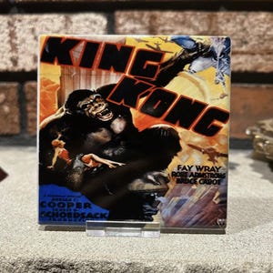 May include: A square tile with the "King Kong" movie poster. The image depicts a giant gorilla, aeroplanes, and the film's title in large letters. The tile is held up by a clear plastic stand.