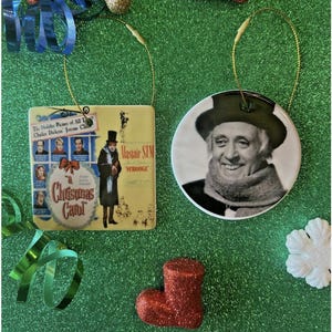 May include: Two Christmas ornaments with gold hanging strings. One is a square ornament with a movie poster design for "A Christmas Carol". The other is a round ornament with a black and white portrait of a man. A red glittery boot and other decorations are also visible.