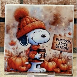 Op de afbeelding: Een vierkante keramische tegel met een cartoonhond die een oranje gebreide muts en trui draagt en een koffiekopje vasthoudt. De scène bevat oranje pompoenen en een houten bord met de tekst PUMPKIN SPICE SEASON in zwarte hoofdletters.