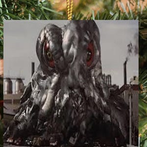 May include: A square-shaped hanging ornament featuring a graphic of a dark, metallic, sludge-like creature with two large, prominent red eyes. The creature is positioned in front of an industrial landscape with smokestacks and factory structures.