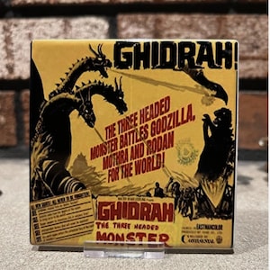 May include: A square, vintage-style print featuring the text "GHIDRAH!" and "THE THREE HEADED MONSTER". The image depicts a three-headed dragon and other monster silhouettes in black against a yellow background. The print is held in a clear stand.