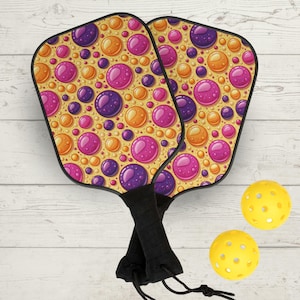 Puede incluir: Dos palas de pickleball con un diseño de burbujas coloridas en rosa, morado y naranja sobre un fondo beige. Las palas tienen mangos negros. También se ven dos pelotas de pickleball amarillas con agujeros.