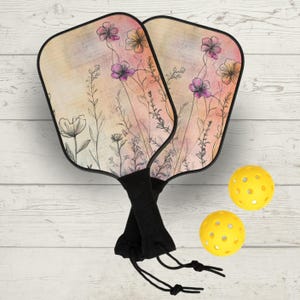 Könnte beinhalten: Zwei Pickleball-Schläger mit einem Blumendesign in Rosa, Lila und Gelb. Die Schläger haben schwarze Griffe und werden mit zwei gelben, perforierten Pickleballs kombiniert. Der Hintergrund ist eine helle Holzmaserung.