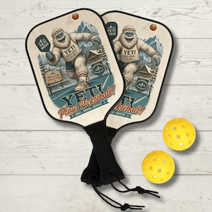 May include: Two wooden pickleball paddles with a cartoon yeti graphic, the text "YETI Plays Pickleball!" and two yellow pickleballs. The paddles have black handles and the image includes the text "YETI Smash" and "High Altitude Good Attitude".