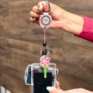 May include: A retractable badge reel with a pink cord, black connector, and a clear ID holder. A flower-shaped key is attached to the holder. The reel is held by a person.