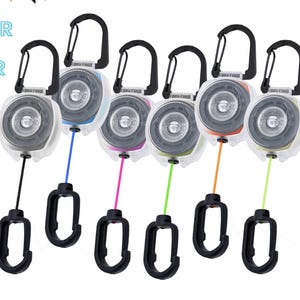 May include: A collection of KEY-BAK POD KEEPER PRO keychains with black carabiner clips and colored cords. The keychains have a clear casing and a retractable cord. The text "NEVER LOSE YOUR EDC" is visible.