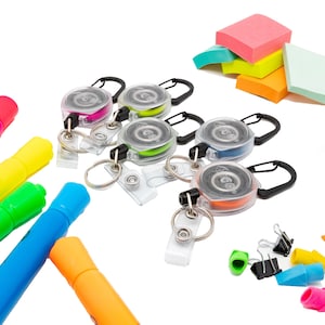 May include: A collection of colorful retractable badge holders with black carabiner clips, along with a variety of office supplies. The badge holders feature pink, green, blue, and orange cords. Also visible are sticky notes and colorful markers.