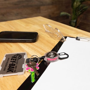 May include: A KEY-BAK retractable key reel with a black carabiner clip, a clear plastic case, and a pink center. A set of keys with a flower-shaped keycap and a phone are also visible on a wooden surface.