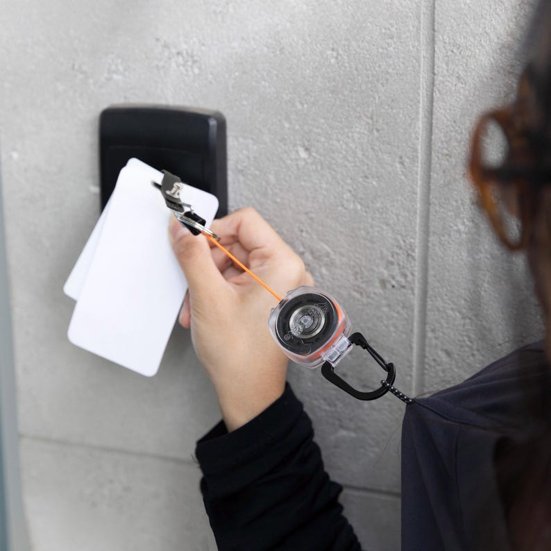 May include: A retractable badge reel with an orange cord and a black carabiner clip. The reel is attached to a person's clothing. The person is holding two white ID cards near a black access panel.