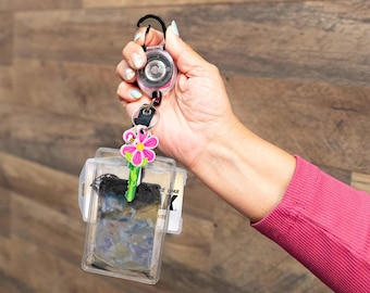 May include: A retractable badge reel with a clear ID holder. The reel is pink and black, and the ID holder is clear. A decorative flower key chain with a pink flower and green stem is attached to the ID holder.