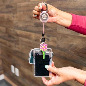 Może przedstawiać: Wysuwana rolka na identyfikator z różowym sznurkiem i przezroczystym uchwytem na identyfikator. Uchwyt na identyfikator zawiera czarną kartę i klucz w kształcie kwiatu. Rolka jest trzymana przez czarny klips i różowo-czarne zapięcie.