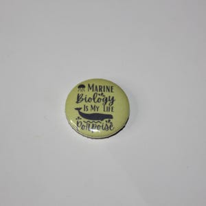 May include: A circular, pale green badge featuring the words "MARINE BIOLOGY IS MY LIFE" in black, with a black whale illustration. The badge also includes the word "porpoise" in black.