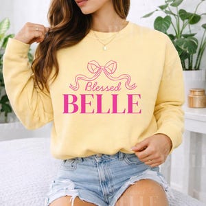 Puede incluir: Sudadera amarilla clara con la inscripción rosa "Blessed BELLE" adornada con un lazo. La modelo lleva un pantalón corto vaquero y joyas doradas. La sudadera es de estilo informal.