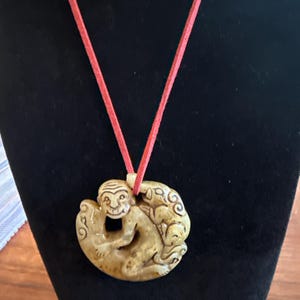May include: A tan-colored pendant necklace featuring a carved monkey design. The pendant is suspended from a red cord. The monkey is depicted in a seated position within a circular frame, with intricate details.