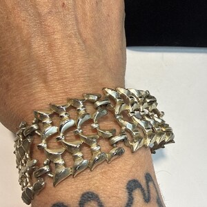 May include: A silver-toned metal bracelet with a textured, articulated design. The bracelet is wide, covering a significant portion of the wrist. The links have a repeating pattern of curved shapes, creating a bold, eye-catching accessory.