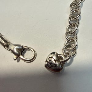 Puede incluir: Pulsera de cadena plateada con un dije en forma de coraz&oacute;n. El dije tiene un dise&ntilde;o grabado y est&aacute; conectado a una cadena de eslabones ovalados. La pulsera cuenta con un cierre de mosquet&oacute;n.