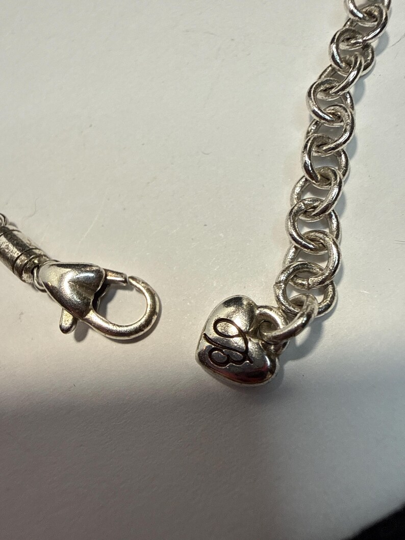 Puede incluir: Pulsera de cadena plateada con un dije en forma de coraz&oacute;n. El dije tiene un dise&ntilde;o grabado y est&aacute; conectado a una cadena de eslabones ovalados. La pulsera cuenta con un cierre de mosquet&oacute;n.