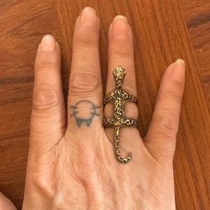 May include: A gold-colored snake ring with a detailed, textured design. The snake's head is at the top, with the body wrapping around the finger and the tail extending down. The ring is worn on a hand with a small tattoo.